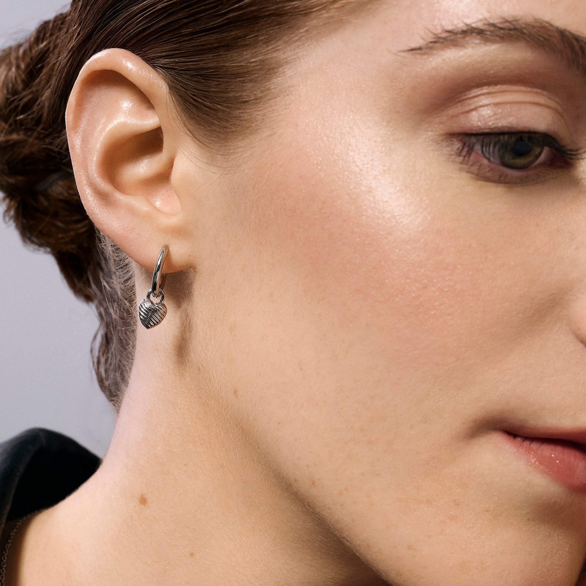 Silver stainless steel hoop earrings with ribbed heart charm worn on ear, romantic minimalist jewelry, hypoallergenic non-tarnish durable earrings, elegant everyday fashion jewelry.