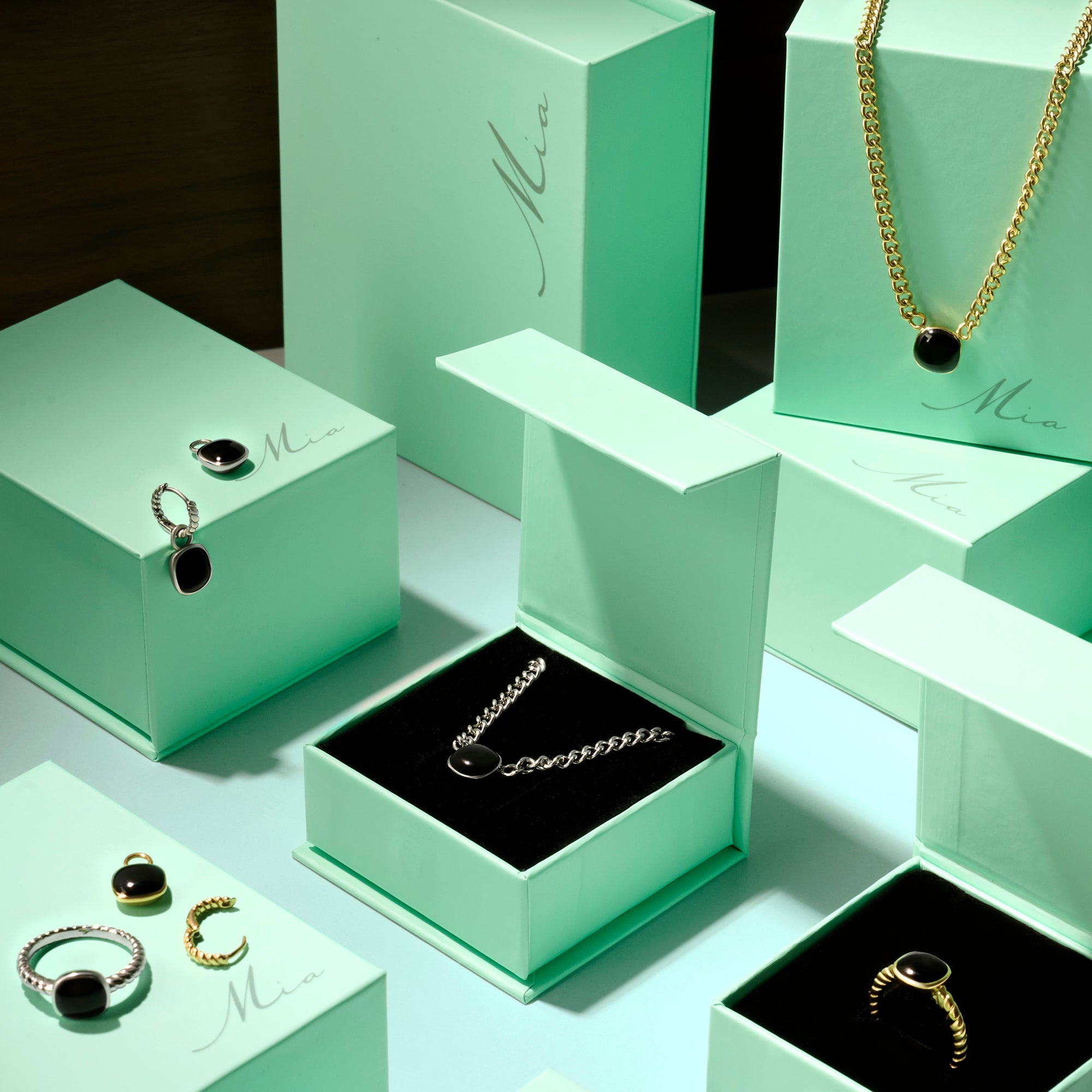 Black gemstone jewelry collection in stainless steel featuring pendant necklaces, rings and earrings displayed in mint green Mia jewelry boxes, elegant modern jewelry with polished black stones.